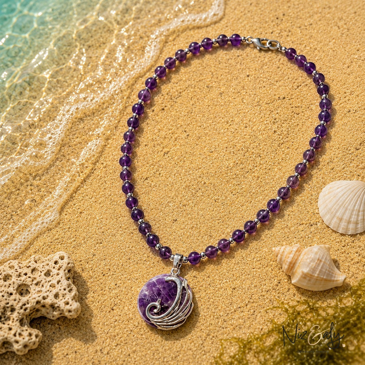 Niz & Aly amethyst beaded necklace with a silver wave pendant resting on sandy beach shore with seashells