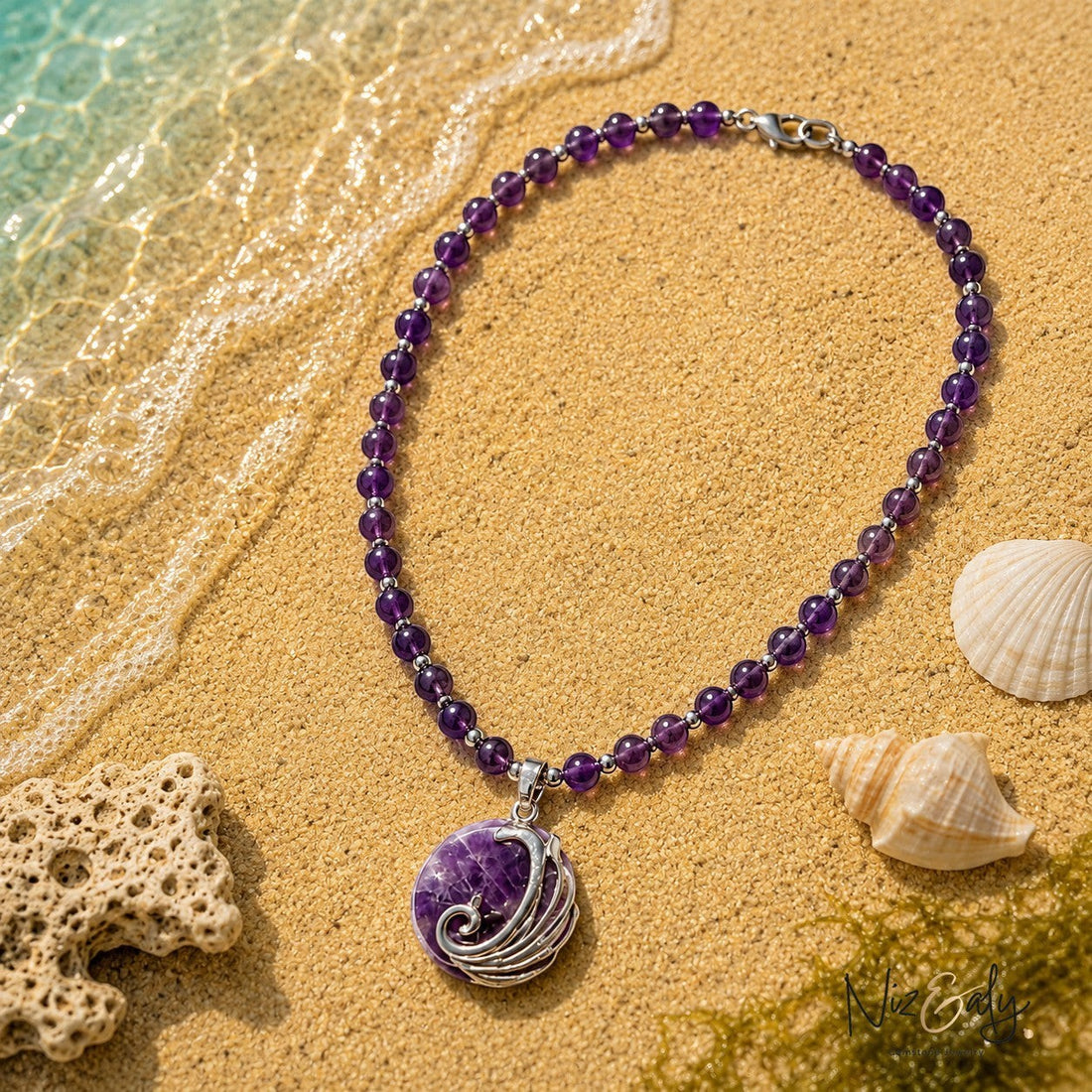 Niz & Aly amethyst beaded necklace with a silver wave pendant resting on sandy beach shore with seashells
