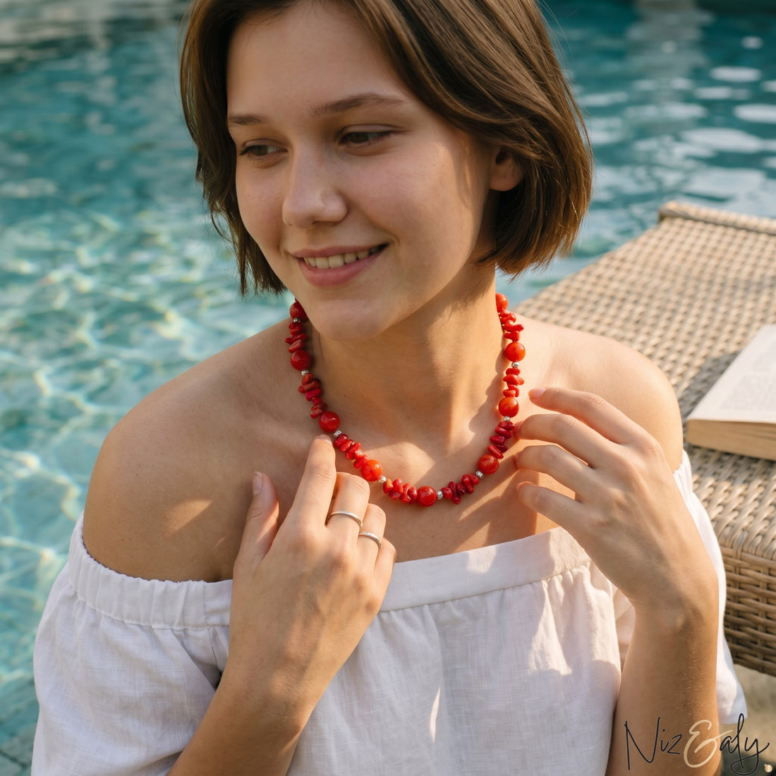 Niz & Aly coral and carnelian bead necklace worn by a woman in a white off-shoulder top by a swimming pool.