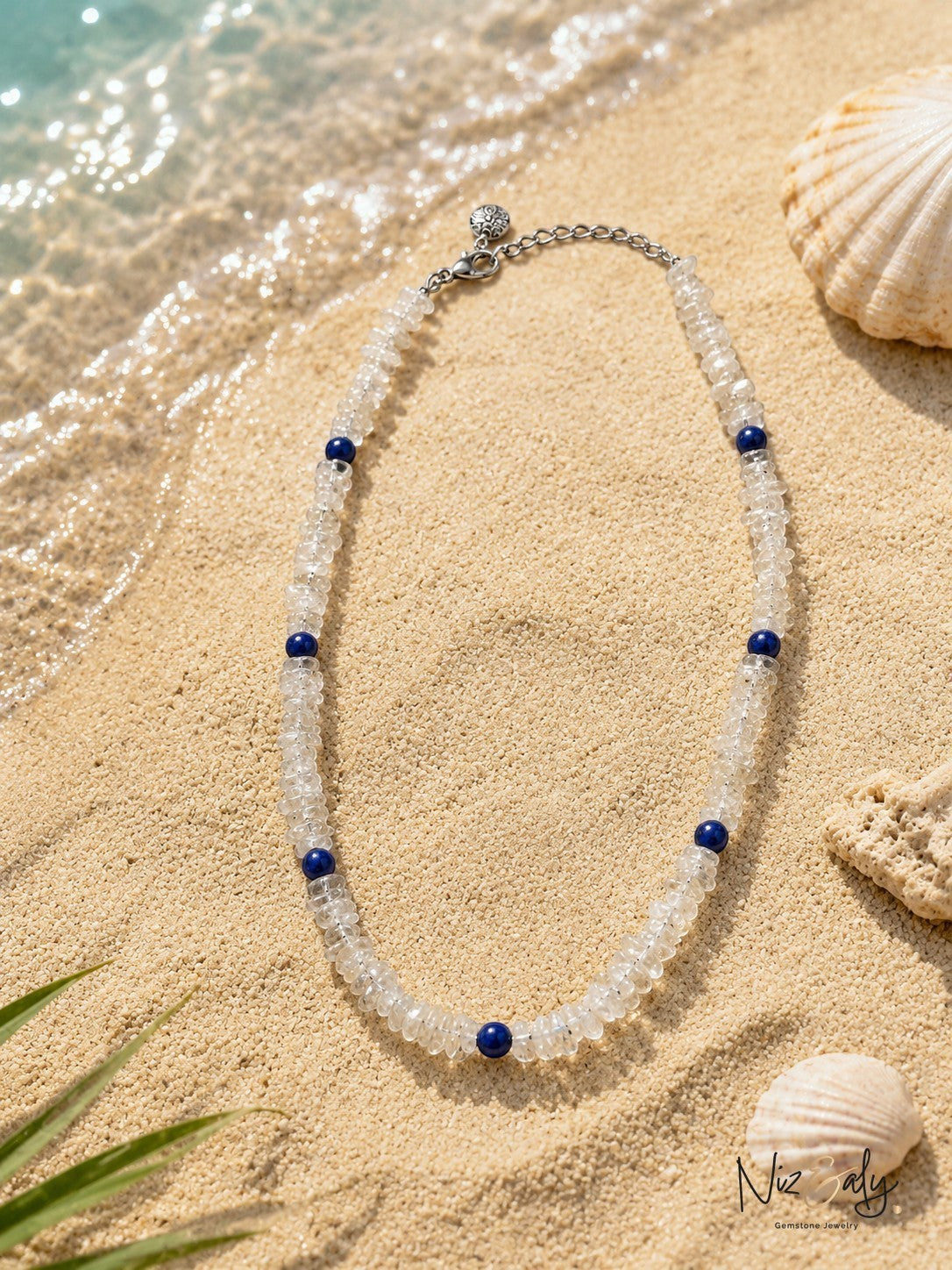 Niz & Aly minimalist necklace featuring clear crystal beads and deep blue lapis lazuli accents on sandy beach shore