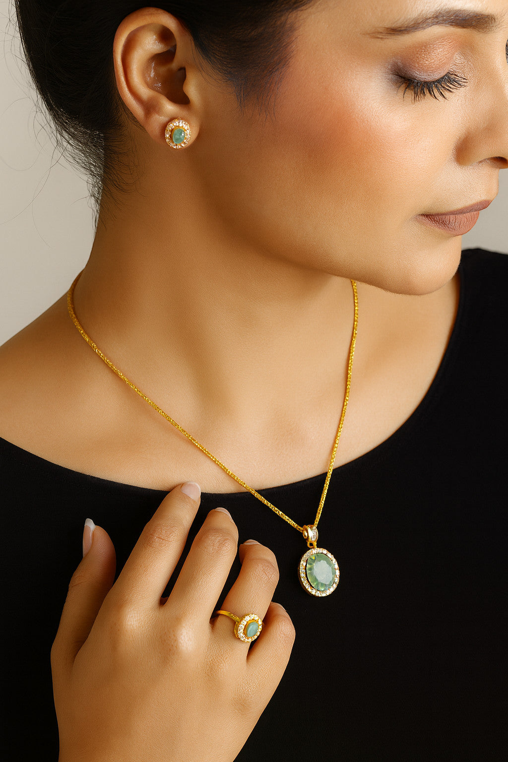 Model wearing Niz & Aly gold-plated emerald halo pendant necklace, matching stud earrings, and ring on black top.