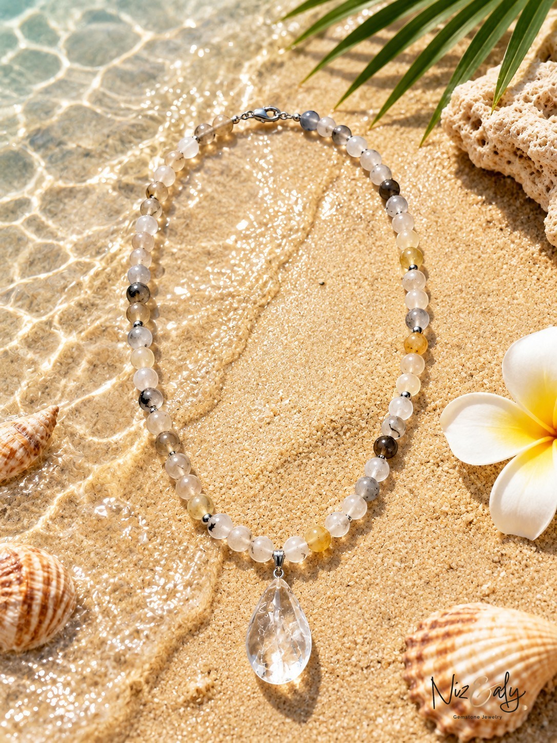 Niz & Aly necklace with natural gemstone beads and a clear teardrop pendant resting on a sunny beach with seashells.