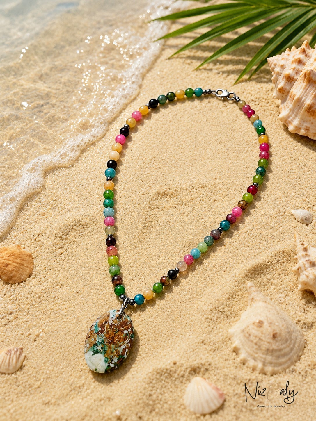 Niz & Aly multi-colored gemstone bead necklace with a large patterned pendant resting on sandy beach shore