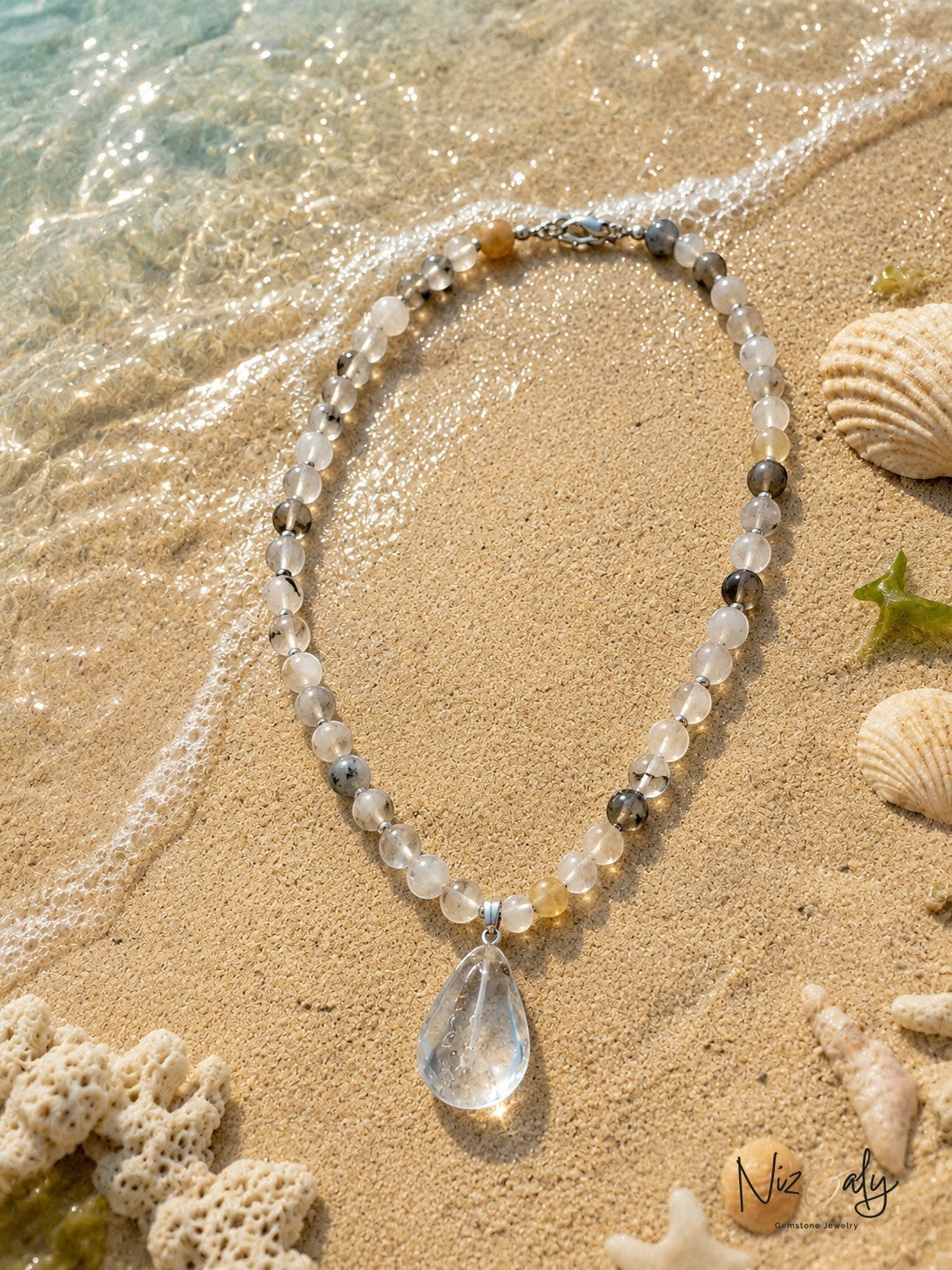 Niz & Aly natural gemstone bead necklace with a clear teardrop pendant resting on sandy beach shore with seashells.