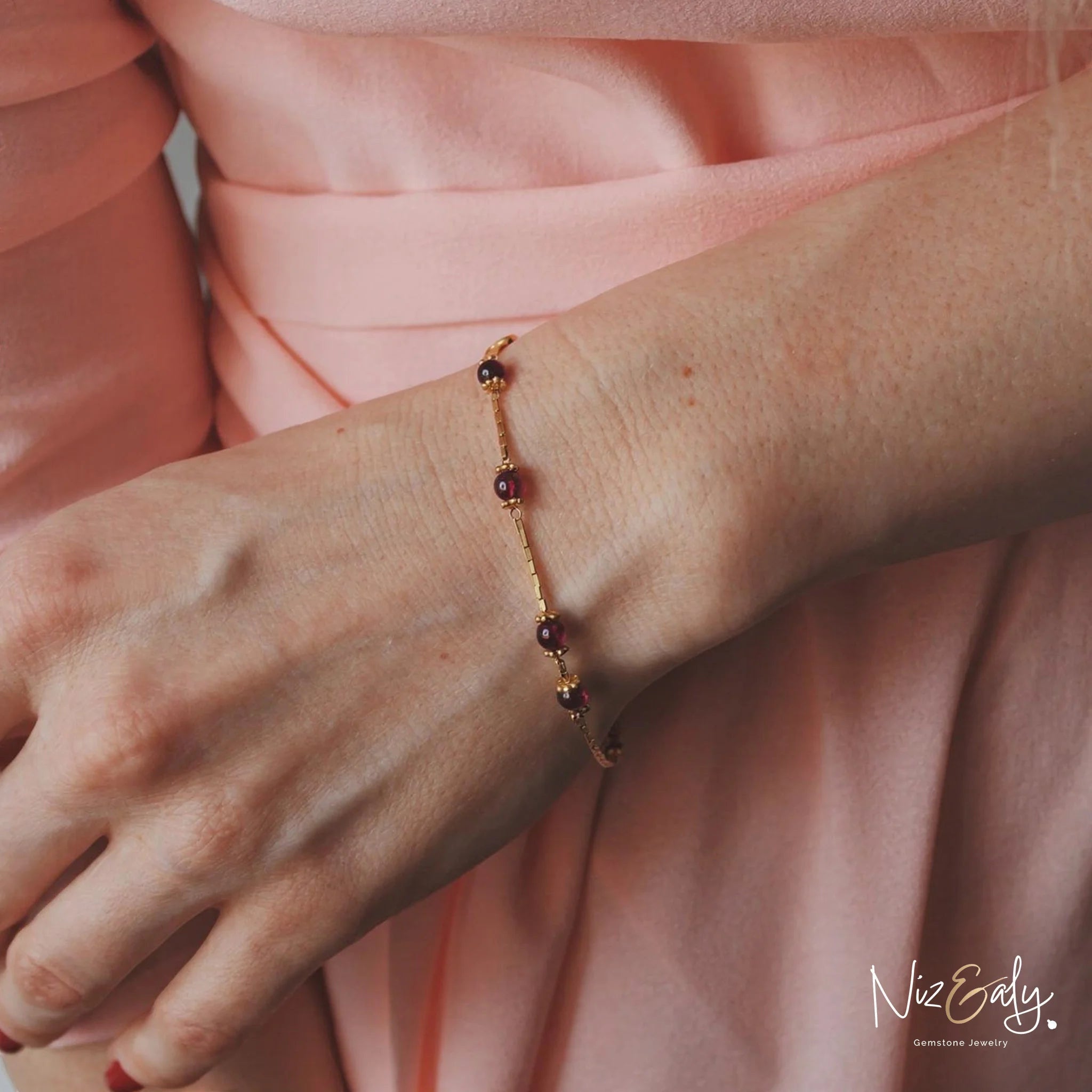 Niz & Aly dainty gold bracelet featuring natural burgundy garnet beads worn on a woman's wrist with a pink sleeve.