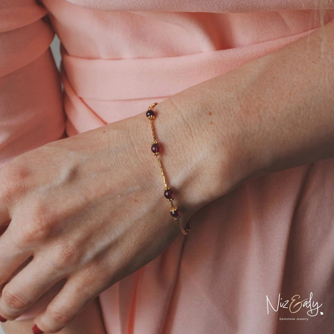 Niz & Aly dainty gold bracelet featuring natural burgundy garnet beads worn on a woman's wrist with a pink sleeve.