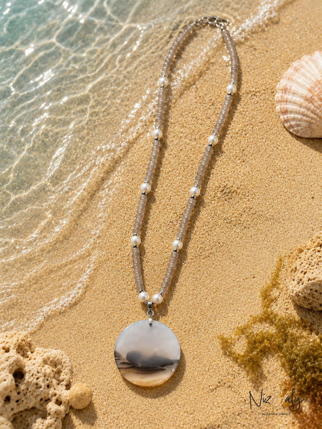 Niz & Aly necklace featuring grey gemstone beads, white pearls, and a large round agate pendant on sandy beach shore.