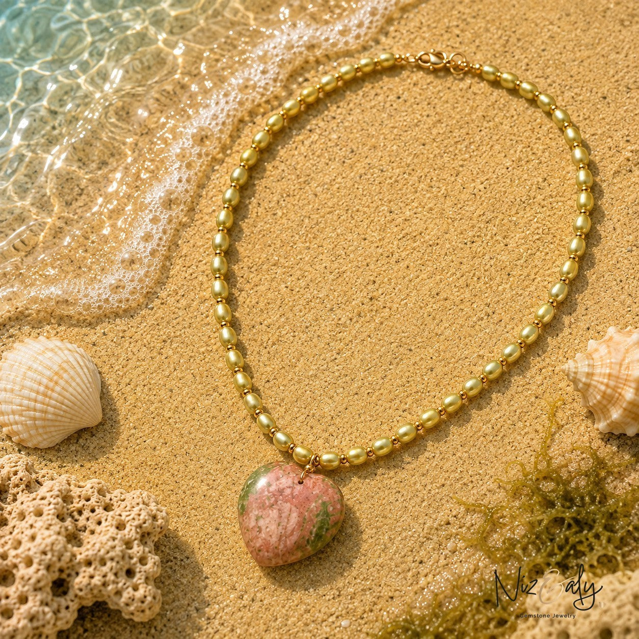 Niz & Aly necklace featuring small gold beads and a heart-shaped pink and green gemstone pendant on sandy beach.