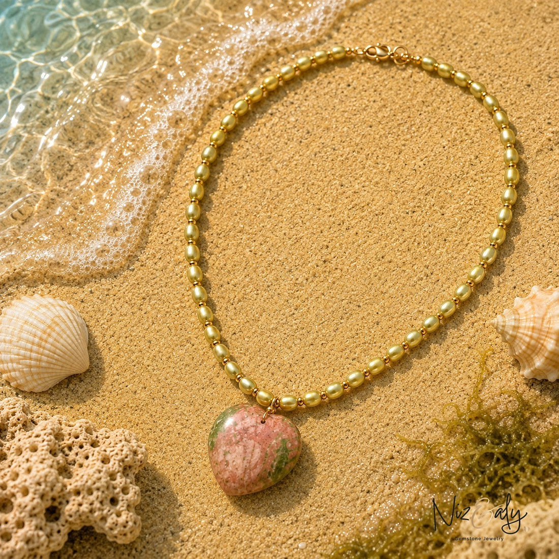 Niz & Aly necklace featuring small gold beads and a heart-shaped pink and green gemstone pendant on sandy beach.