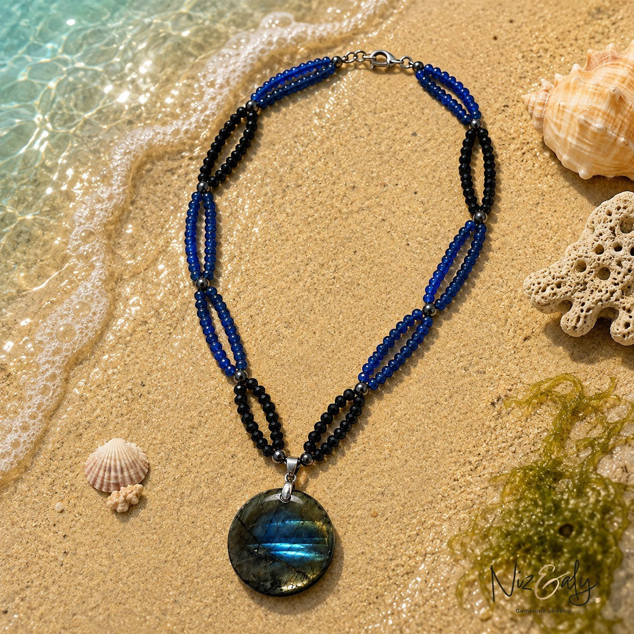 Niz & Aly blue and black beaded gemstone necklace with a large iridescent labradorite pendant on a sandy beach
