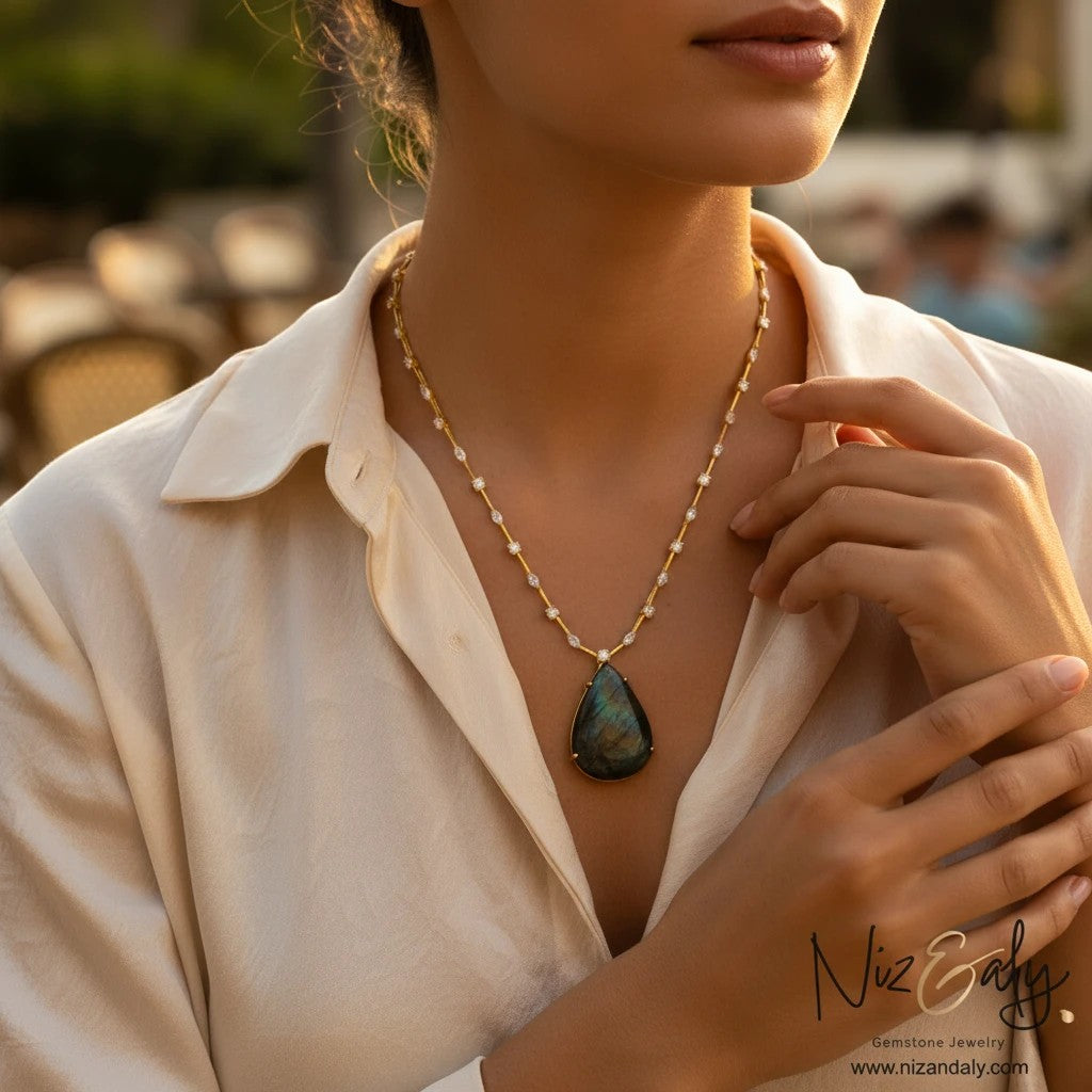 Niz & Aly teardrop labradorite pendant on a gold-toned studded chain worn by a woman in a cream silk shirt