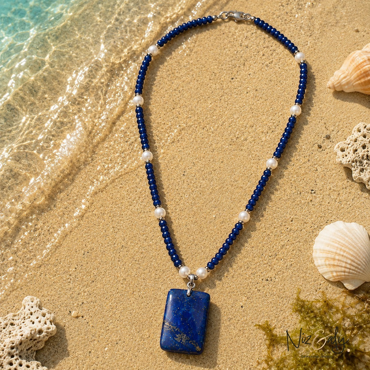 Niz & Aly necklace with blue lapis lazuli beads, white pearls, and a rectangular lapis pendant on sandy beach shore.