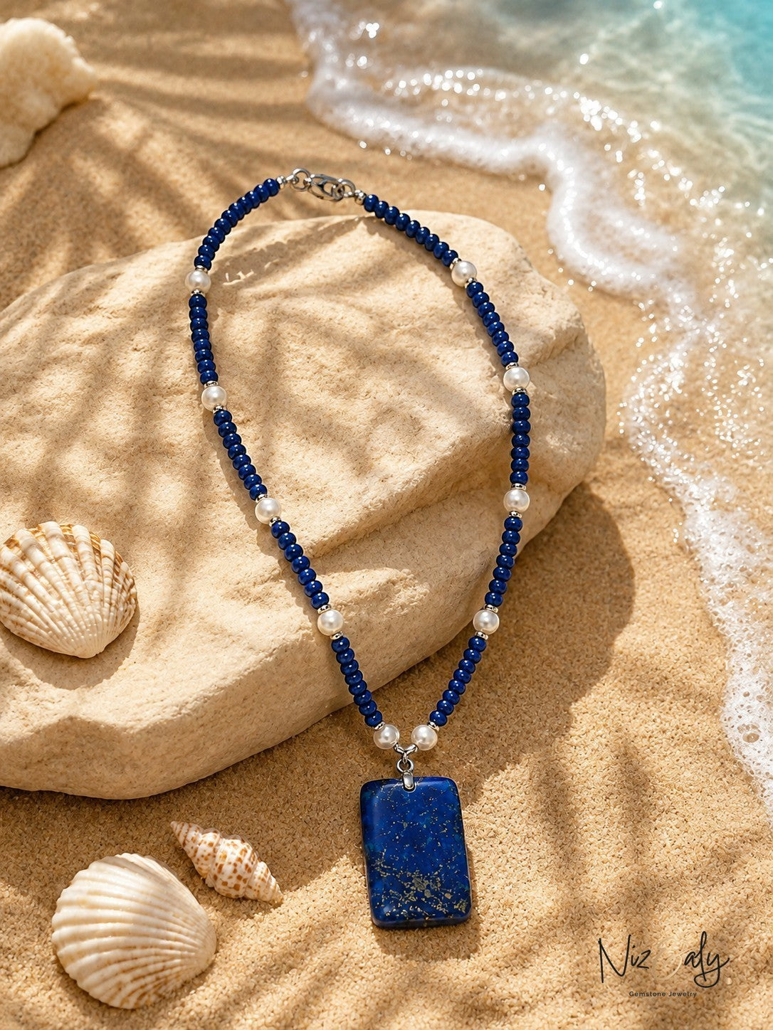 Niz & Aly blue lapis lazuli gemstone and pearl beaded necklace resting on a sandy beach with seashells.