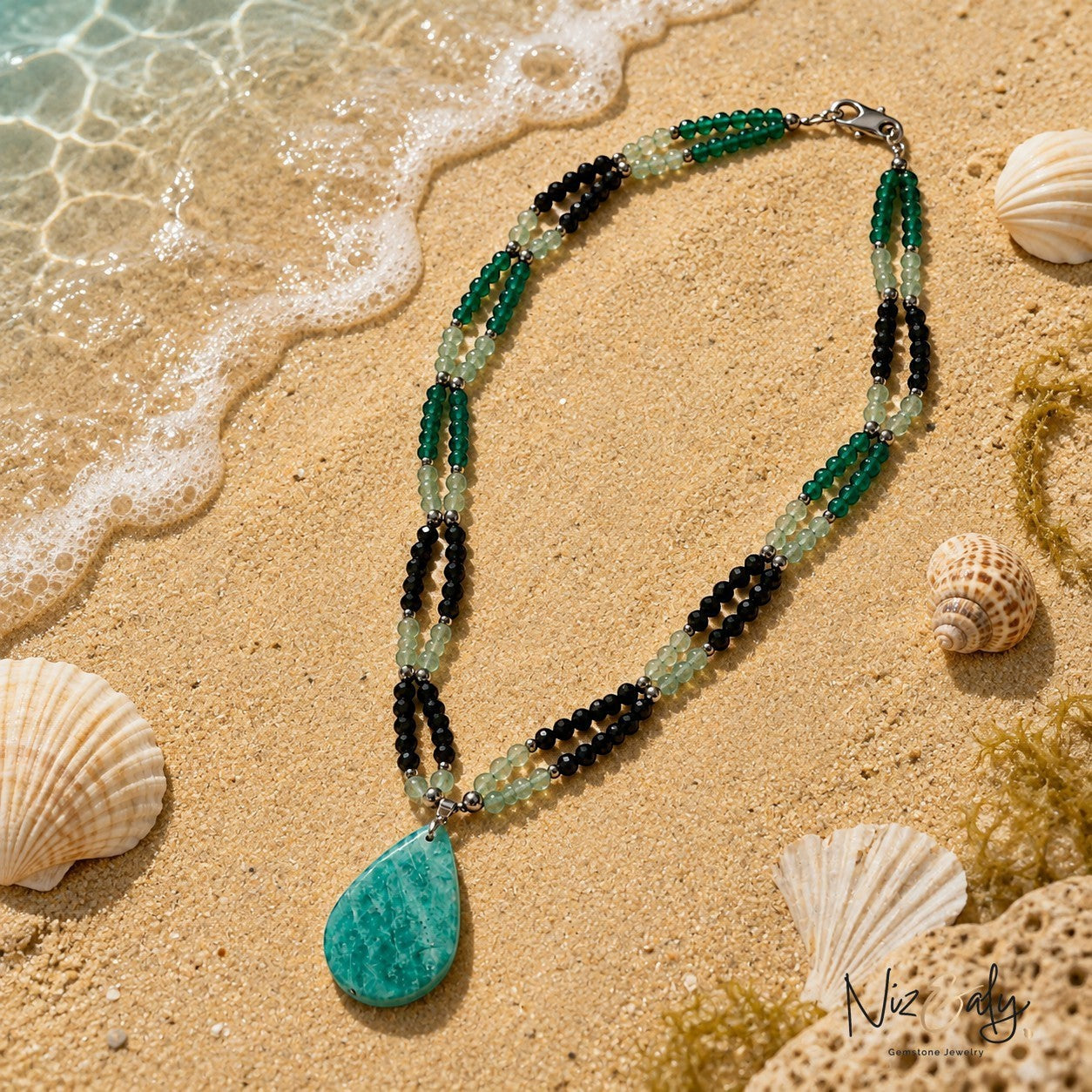 Niz & Aly layered gemstone necklace featuring green and black beads with a large teardrop pendant on sandy beach