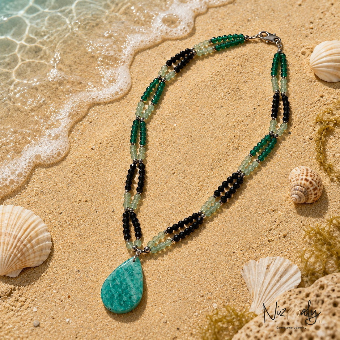 Niz & Aly layered gemstone necklace featuring green and black beads with a large teardrop pendant on sandy beach