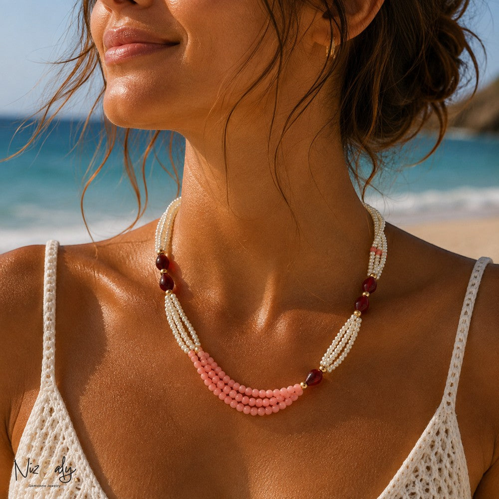 Niz & Aly layered gemstone necklace with white pearls, pink beads, and dark red accents worn on a beach.