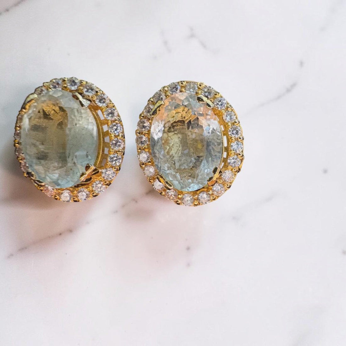 Niz & Aly oval aquamarine halo stud earrings in polished gold with brilliant accent stones on a marble surface.