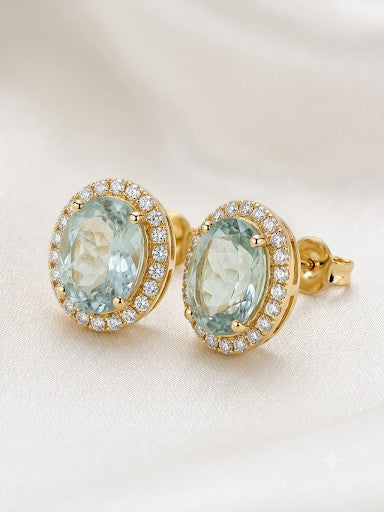 Niz & Aly oval aquamarine halo stud earrings in polished gold with sparkling white accent stones on silk fabric.