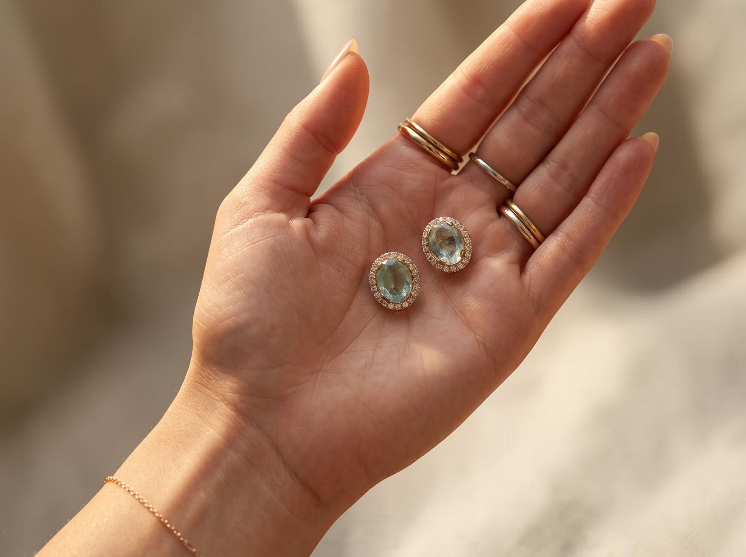 Niz & Aly oval aquamarine halo stud earrings with brilliant stones set in gold, held in a palm.