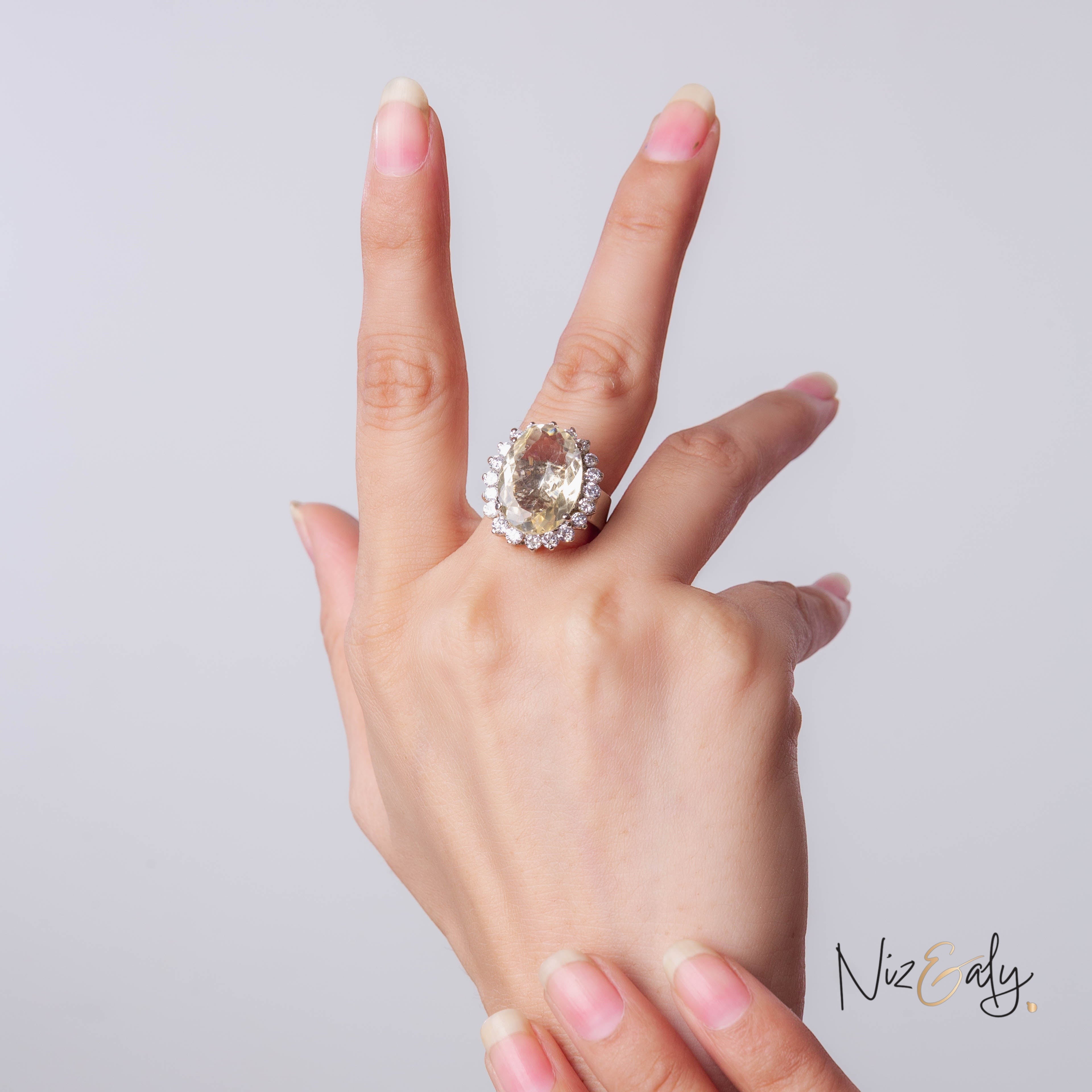 Niz & Aly designer ring featuring a large oval citrine stone surrounded by a halo of sparkling diamonds on a hand.