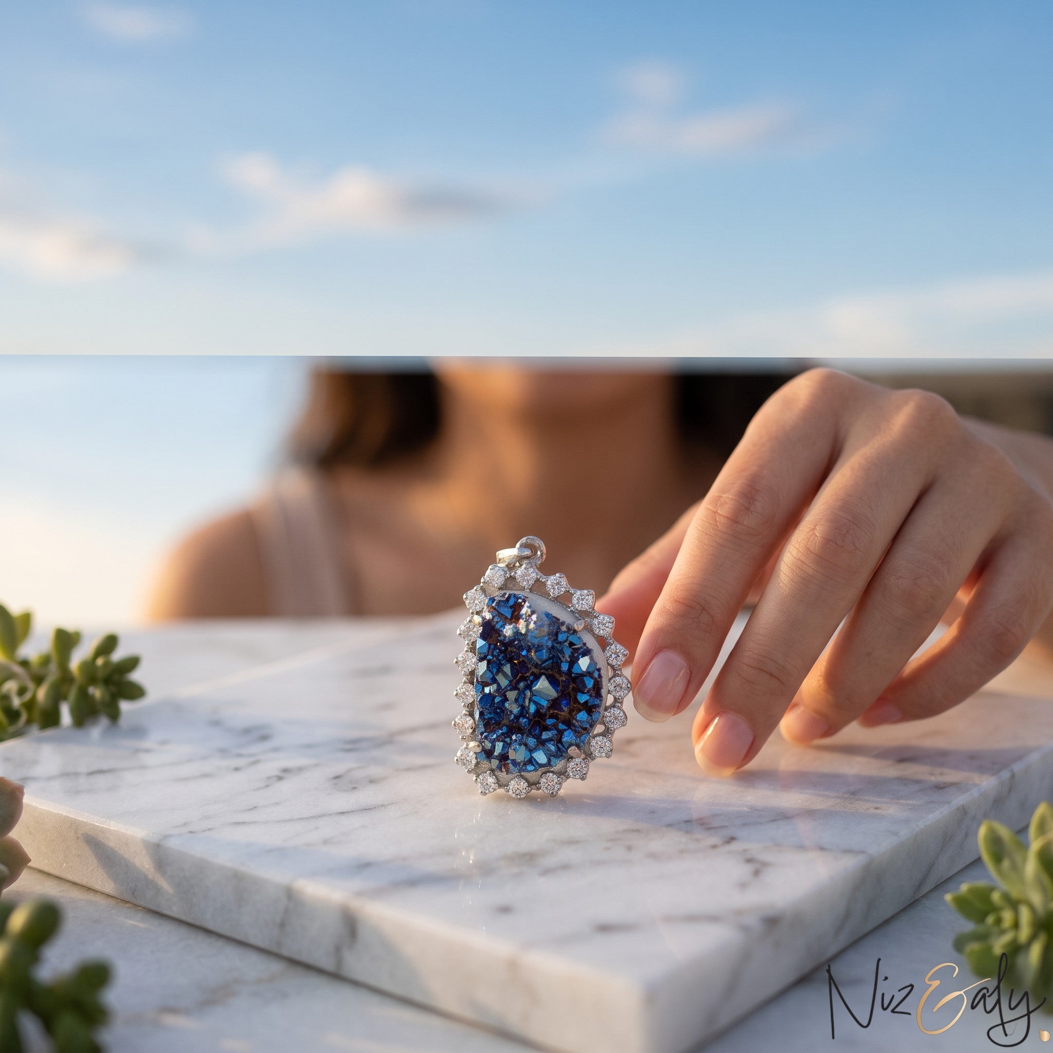 Niz & Aly peacock ore bornite pendant with iridescent blue crystals in a sterling silver setting on marble surface
