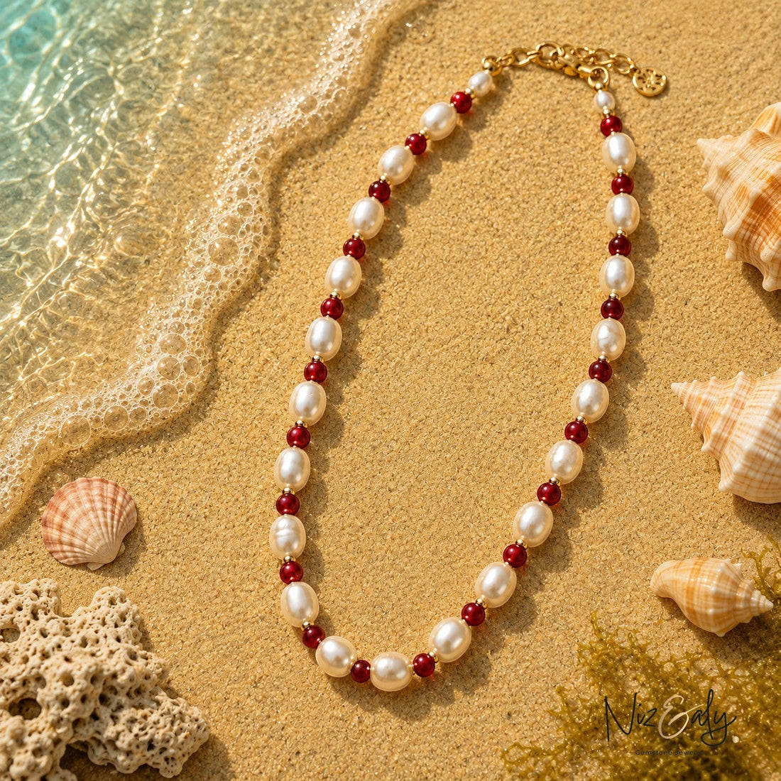 Niz & Aly necklace featuring white freshwater pearls and small red gemstone beads on a sandy beach shore.