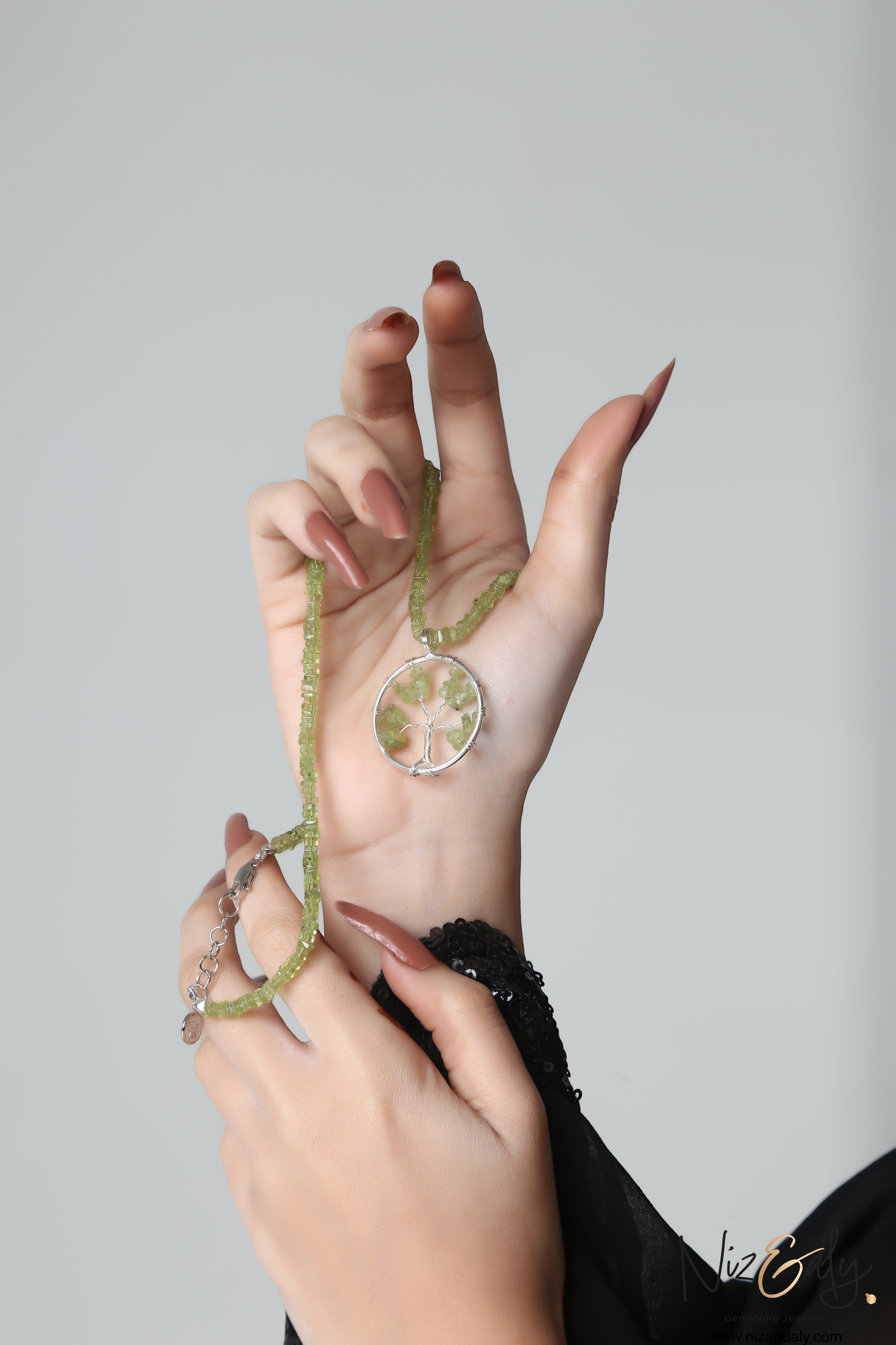 Niz & Aly peridot tree pendant necklace with green heishi-cut gemstone beads held in a hand against a grey background.