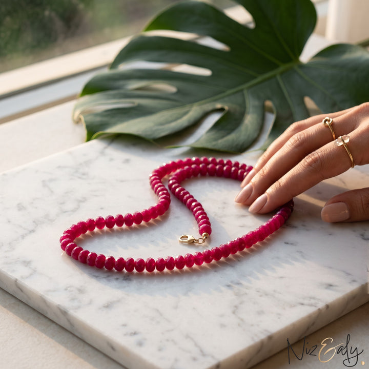 Niz & Aly red jade bead strand necklace with polished scarlet gemstones on a marble surface with a monstera leaf.