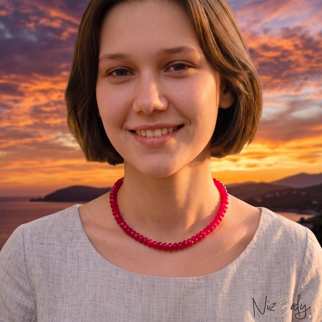 A woman wearing a Niz & Aly scarlet red jade bead strand necklace with a light grey linen top in a sunlit bedroom.