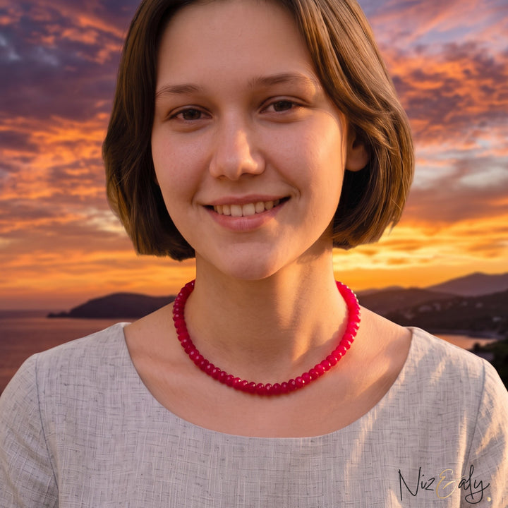 A woman wearing a Niz & Aly scarlet red jade bead strand necklace with a light grey linen top in a sunlit bedroom.