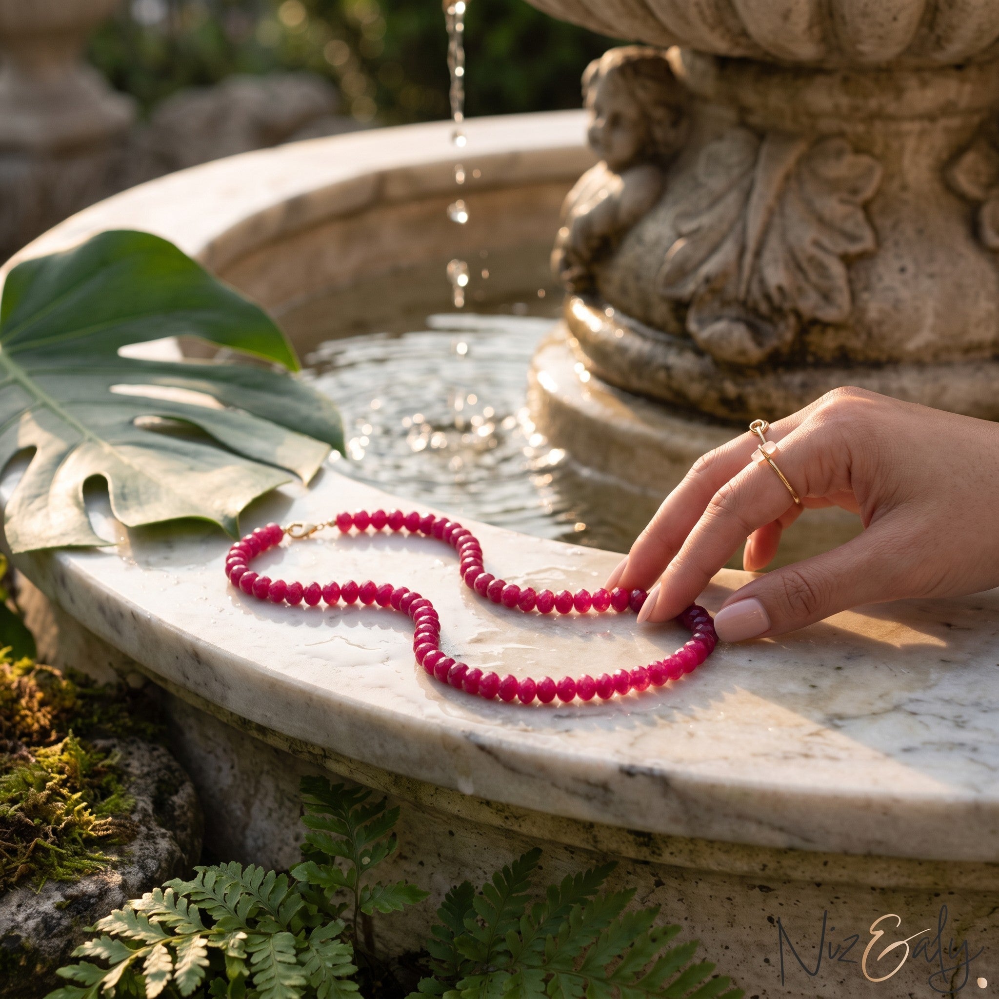 Niz & Aly red jade bead strand necklace resting on a marble fountain edge near a monstera leaf