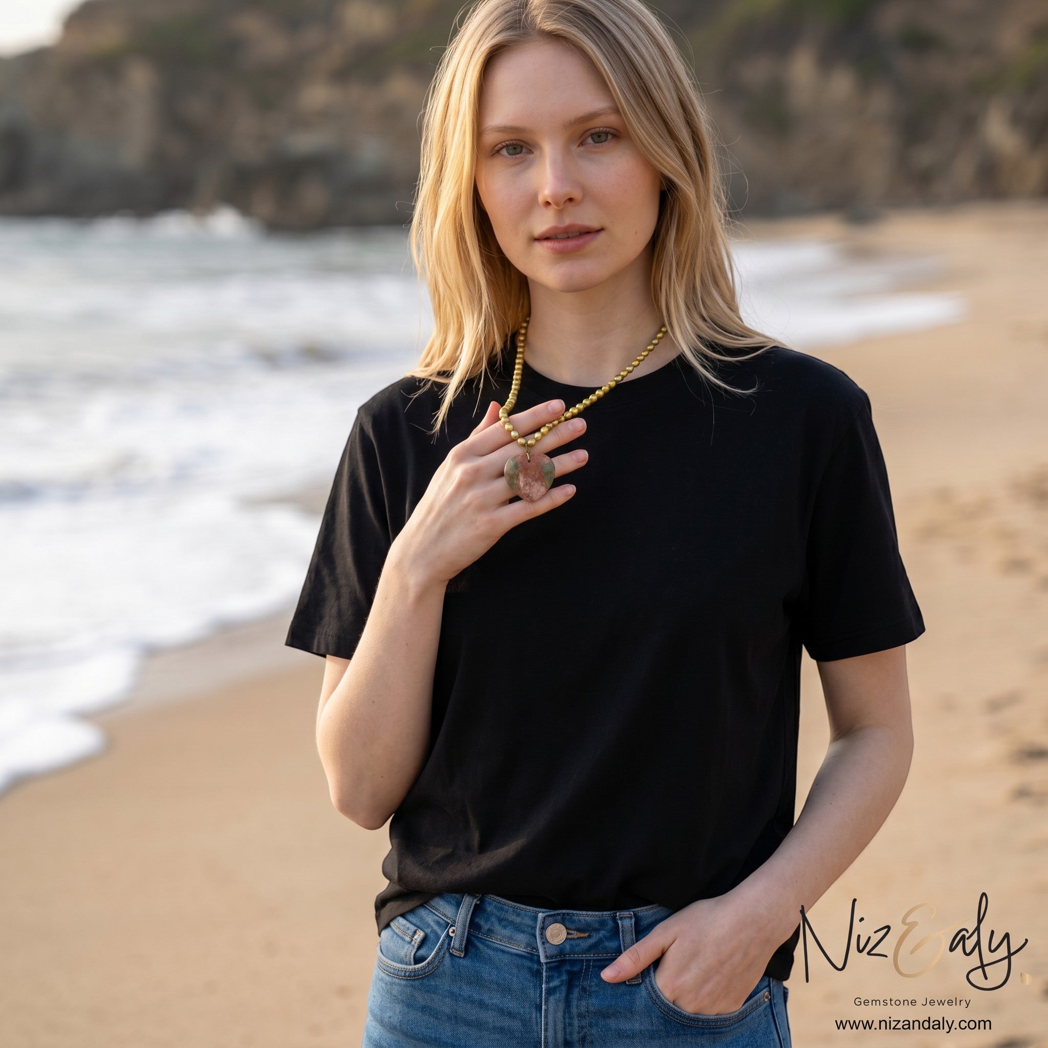 Niz & Aly unakite heart pendant on a golden rice pearl necklace worn by a woman on a beach.