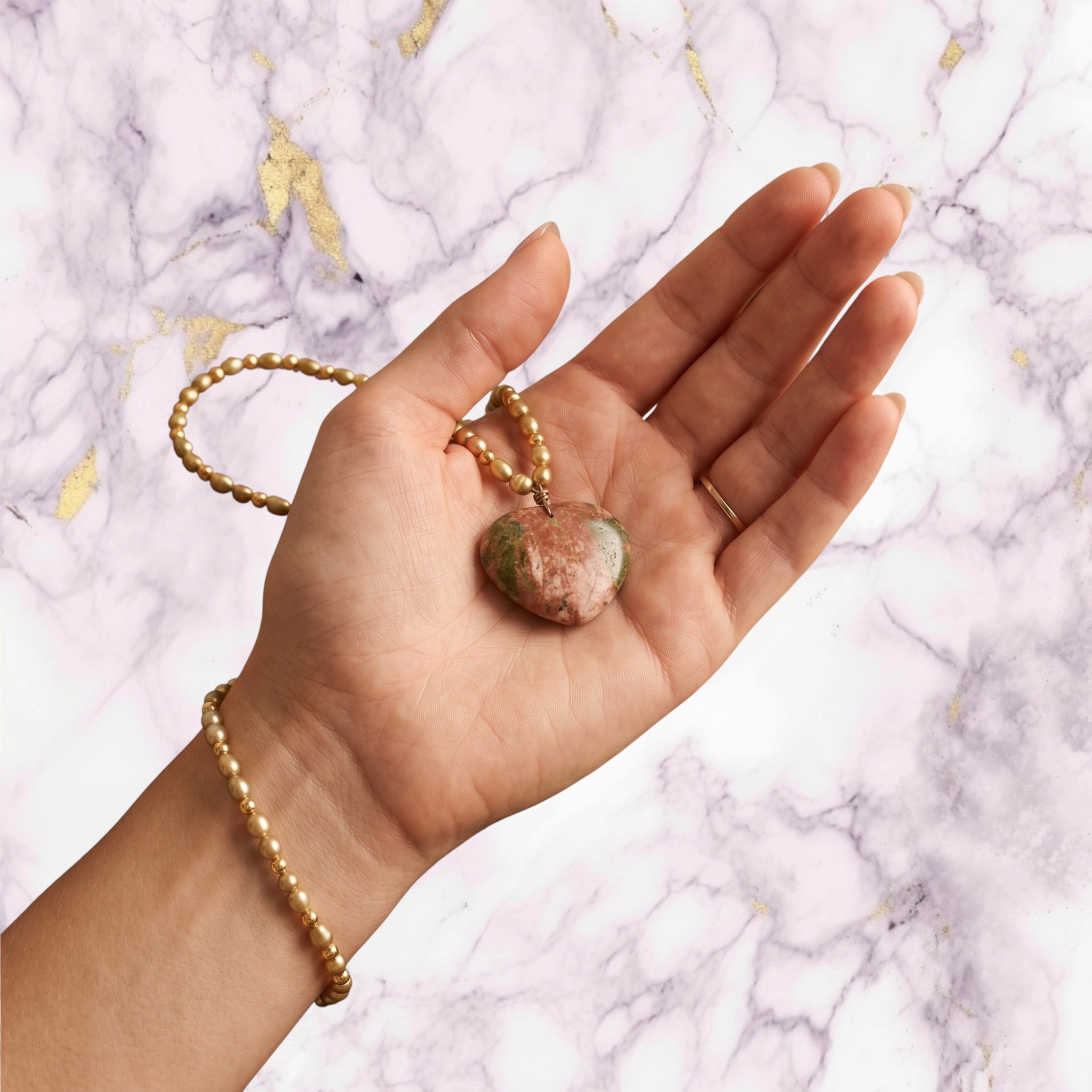 Niz & Aly unakite heart pendant on a golden rice pearl necklace held in a hand against a linen background.